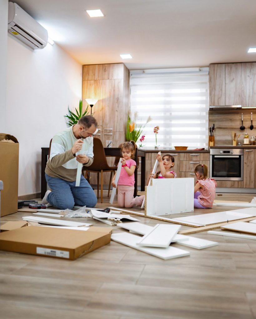 In a modern kitchen, a parent helps three children build furniture, surrounded by boxes and parts. The cheerful atmosphere highlights teamwork and creativity while working together.