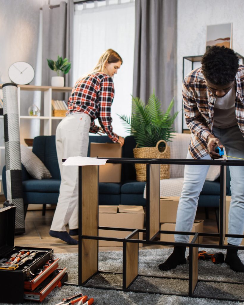 Handsome african american man using tape for measuring length of bookcase at living room. Charming caucasian woman picking up boxes from blue comfortable sofa near. Moving concept.
